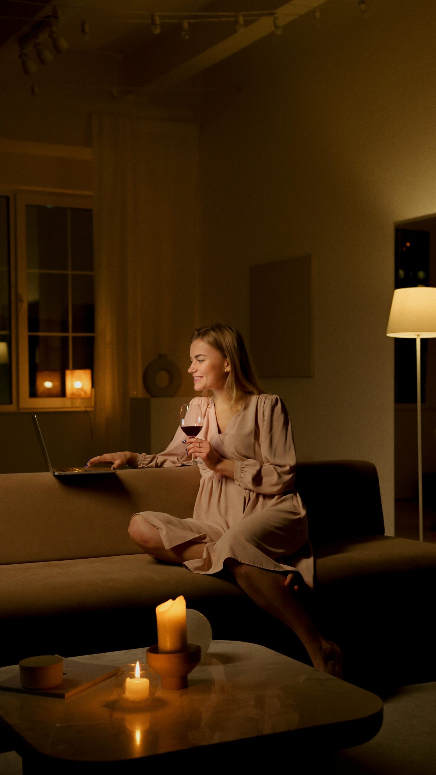 A woman enjoys a cozy evening indoors, sitting on a couch with a laptop and glass of wine.