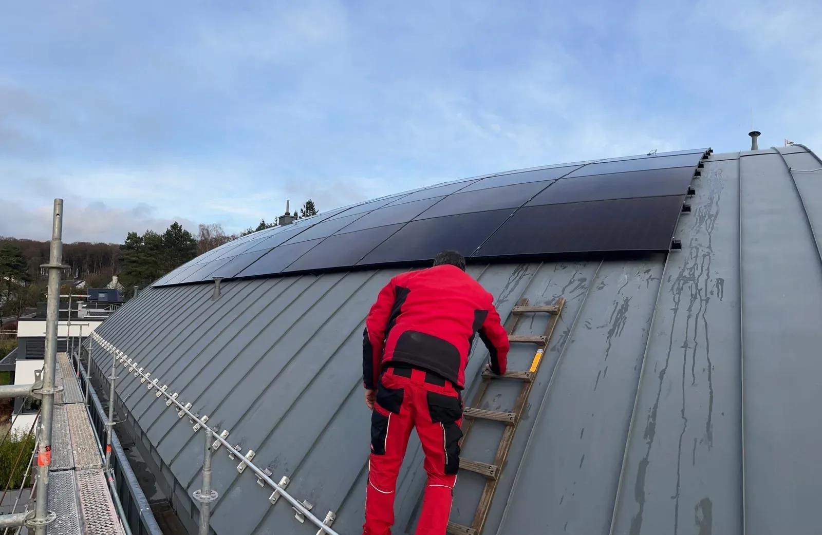 Installateur en tenue de sécurité rouge posant des panneaux solaires photovoltaïques noirs sur une toiture cintrée en zinc sécurisée.
